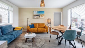 A modern living room with a blue sofa, a mustard-colored couch, a wooden dining table, and a large window with blinds.