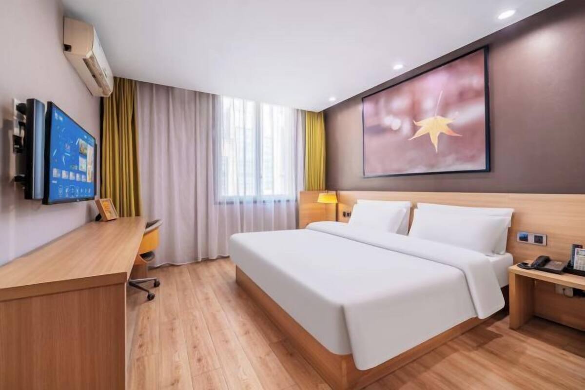 A hotel room with a bed, desk, chair, bedside table, lamp, and a TV mounted on the wall.