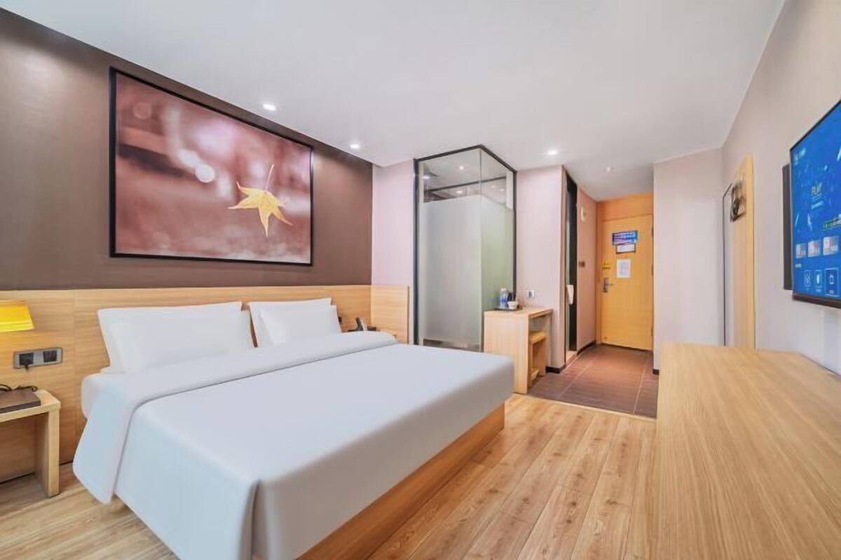 A hotel room with a bed, a nightstand, a wall-mounted TV, and a glass partition.