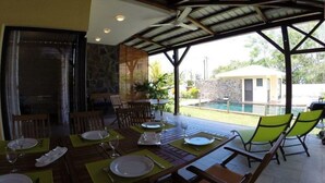 Un área de comedor al aire libre con mesas dispuestas para dos, una piscina al fondo y un muro de piedra.