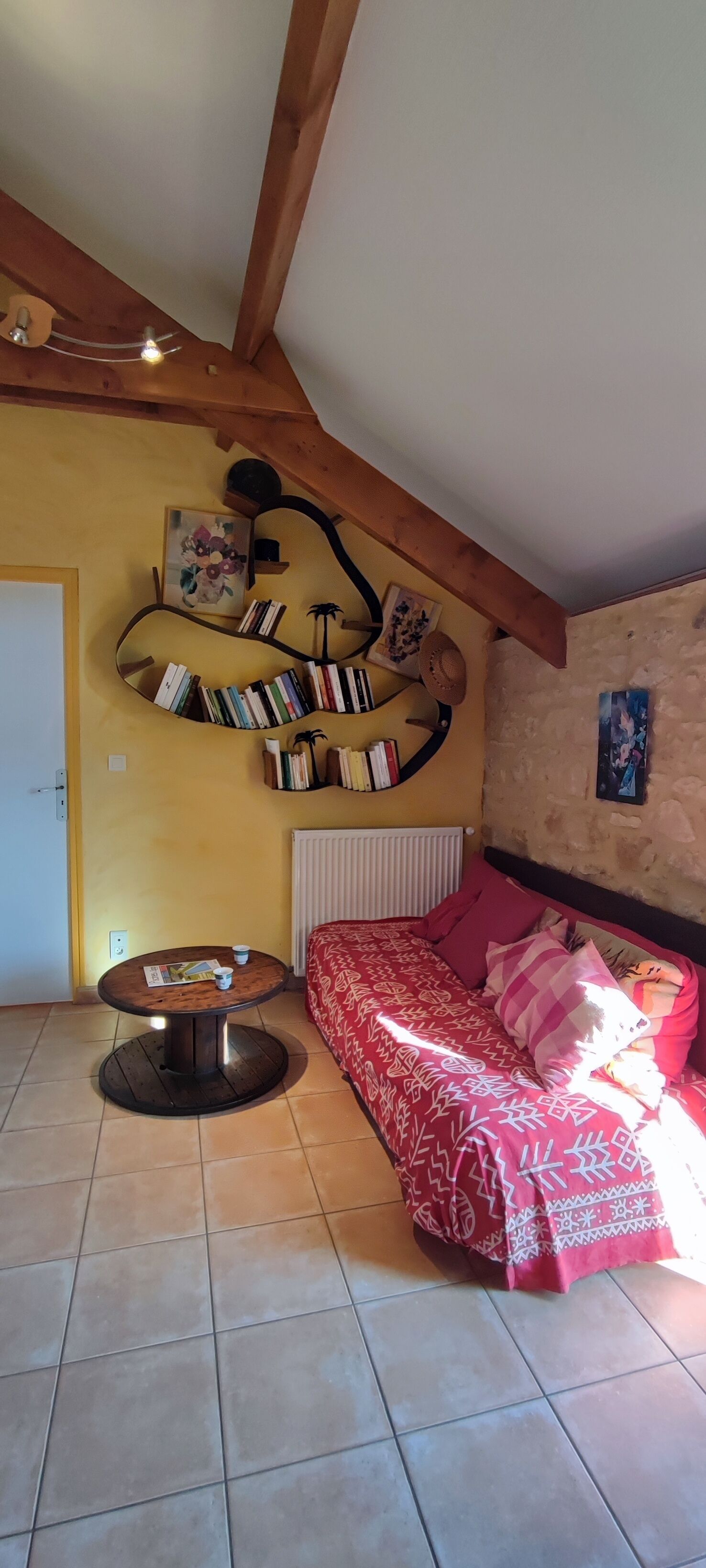 Une pièce chaleureuse avec un canapé rouge à motifs, une table ronde en bois sur laquelle sont posés des magazines et une bibliothèque fixée au mur.