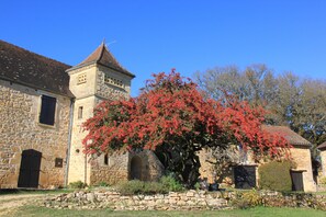 A stone building with a tower and arched windows, surrounded by a stone wall and a tree with red foliage.