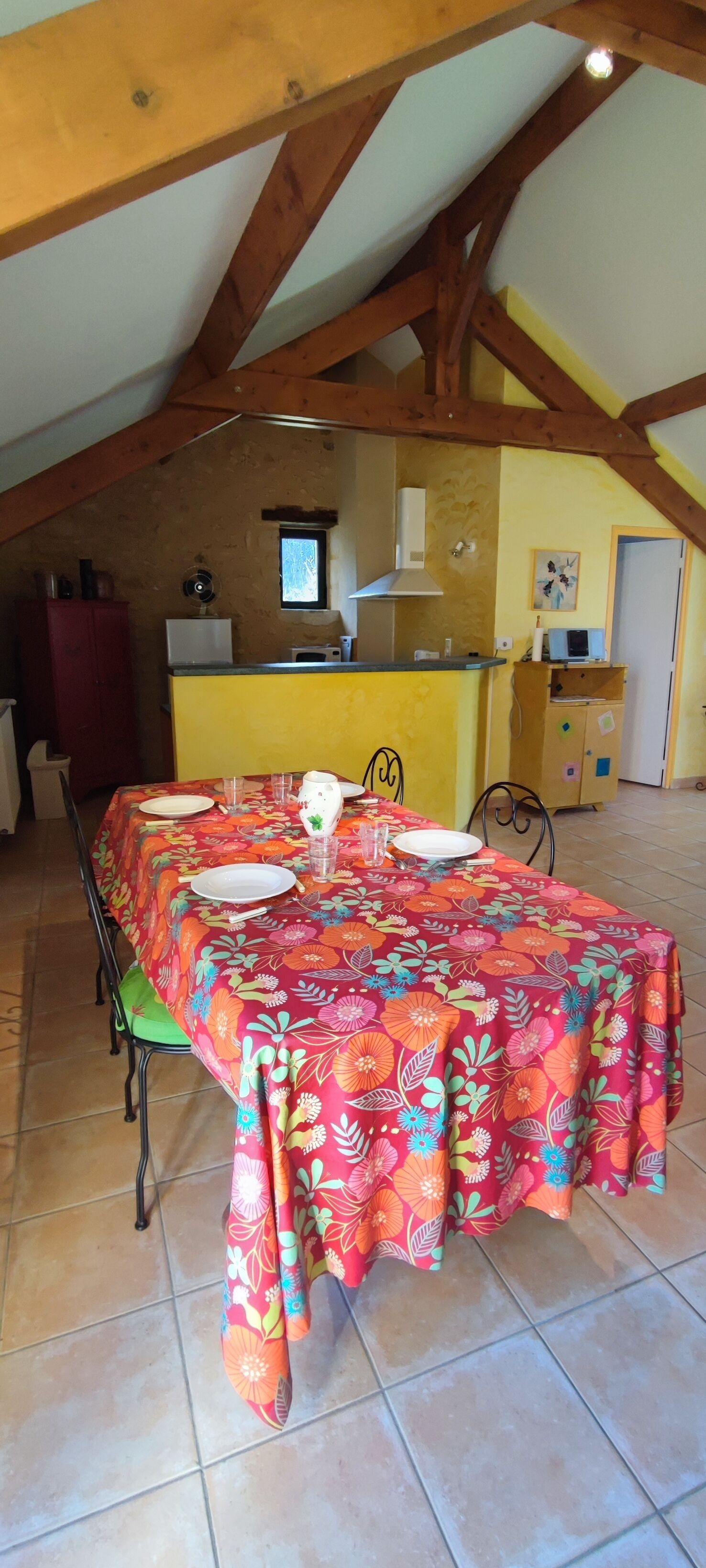A dining area with a table set for four, a kitchen in the background, and a sloped ceiling with wooden beams.