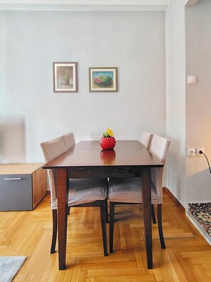 A dining area with a wooden table, cushioned chairs, a red vase with flowers, and two framed pictures on the wall.