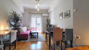 Uma sala de estar com sofá, mesa de jantar, cadeiras, mesa de centro, planta em vaso e televisão.