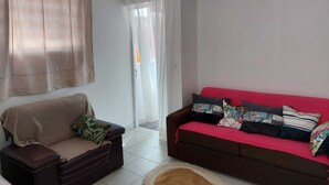 A living room with a beige sofa, a red sofa with colorful cushions, a brown ottoman, and a white curtain. - APPT T2 Cirique (Anses d'Arlet)