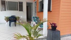 A covered patio area with a dining table and chairs, a potted plant, and a building with orange siding. - APPT T2 Cirique (Anses d'Arlet)