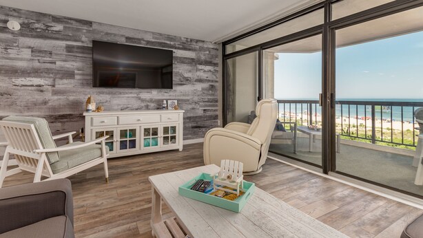 A balcony with a view of the beach, a white sofa, a white armchair, a small table with a tray, and a TV mounted on a wood-textured wall. - Direct oceanfront rennovated condo with panoramic views of ocean and bay right on the beach! (Ocean City)