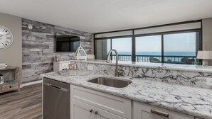 A modern kitchen with a marble countertop, a built-in dishwasher, and a large window overlooking the sea. - Direct oceanfront rennovated condo with panoramic views of ocean and bay right on the beach! (Ocean City)