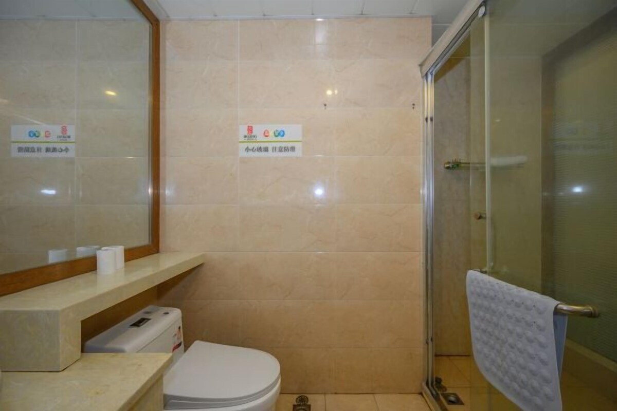 Baño con inodoro, lavamanos y zona de ducha. Hay señalizaciones en la pared y una toalla colgando de la puerta de la ducha.