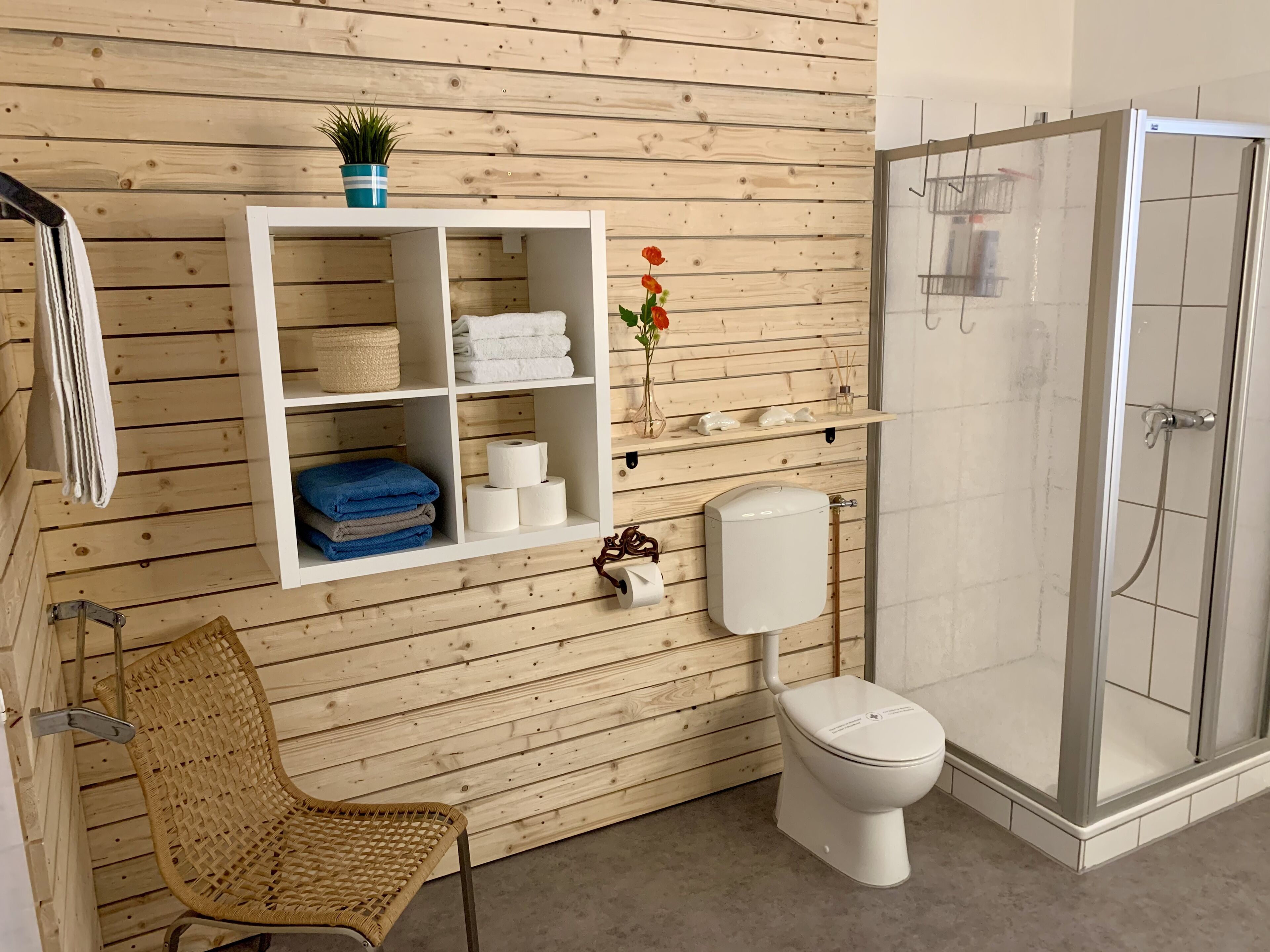 Ein Badezimmer mit Holzwand, einem weißen Ablagebrett mit Handtüchern, einer Toilette und einer Duschkabine.