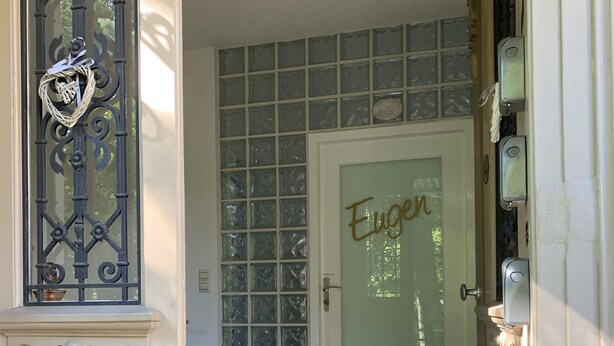 A hallway with a checkered floor, a decorative metal gate, a frosted glass door with the name 'Eugen' on it, and a tiled wall.