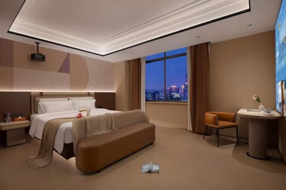 A modern hotel room with a bed, a sofa, a desk, and a view of the cityscape.