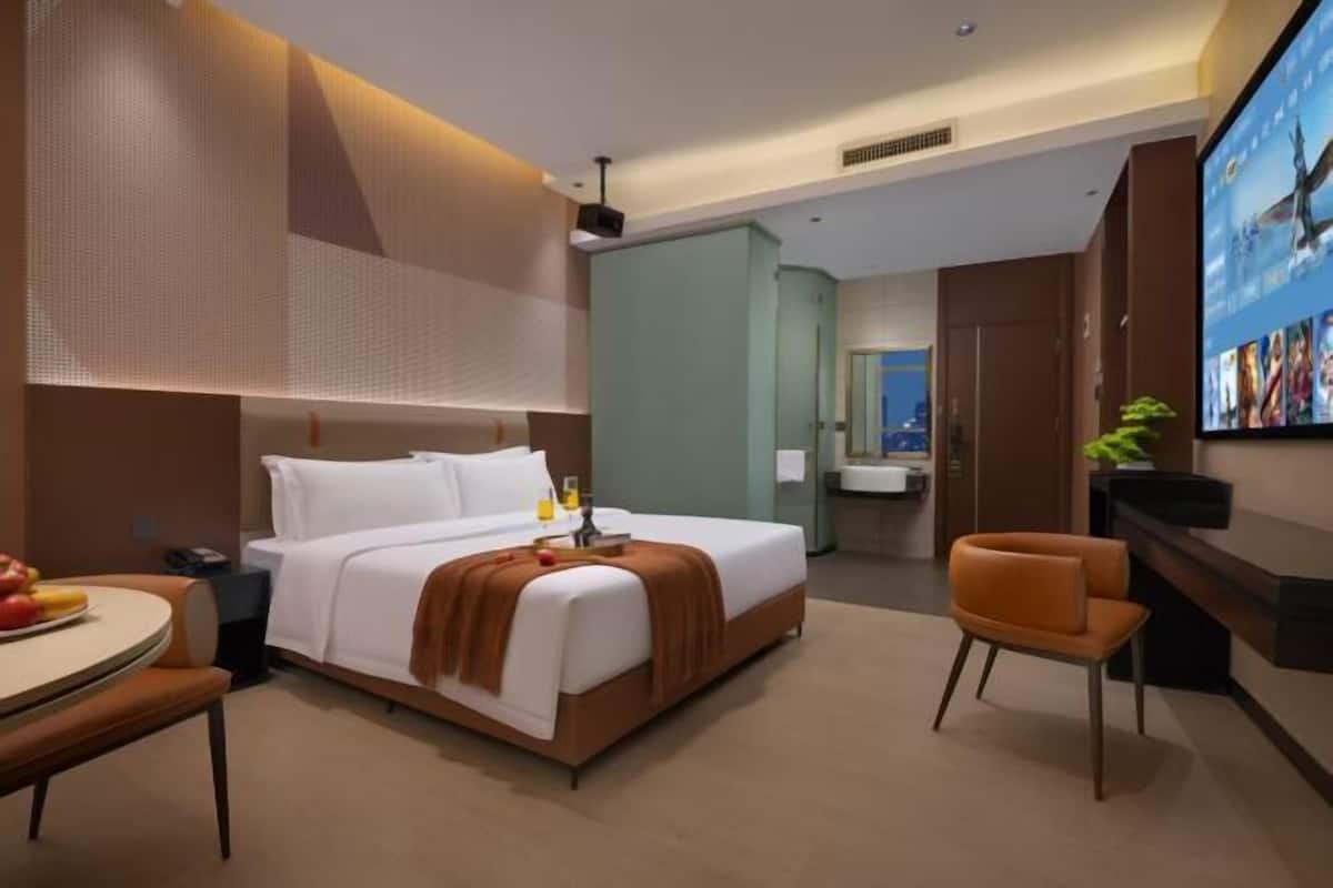A modern hotel room with a bed, a desk, a chair, a television, and a bathroom visible in the background.
