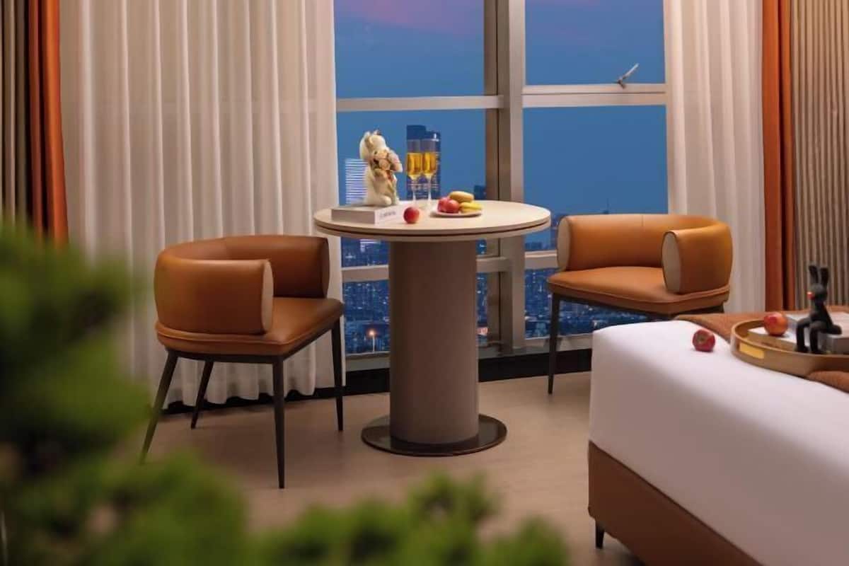 A hotel room with a city view, a round table with fruit and drinks, two leather chairs, and a bed with a tray of apples.