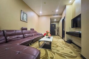 A hotel room with a leather sofa, a small coffee table with flowers, a flat-screen TV mounted on the wall, and a carpeted floor. - Jiangmen Jinxi International Hotel (Wuyi University Lihe Plaza) (Jiangmen)
