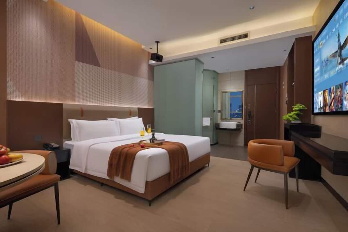 A modern hotel room with a bed, a desk with a chair, a television, and a bathroom visible in the background.