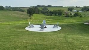 Een rond zitgedeelte buiten, met vier Adirondack-stoelen, omringd door een verzorgd gazon en uitzicht op een landelijk landschap met velden en bomen.