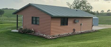 A single-story log cabin with a green metal roof, a small porch, and a clear view of the surrounding grassy landscape.