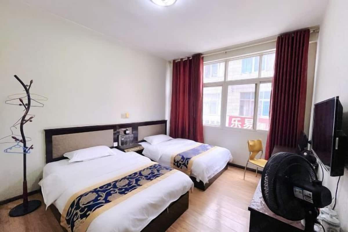 Ein Hotelzimmer mit zwei Betten, einem Standventilator, einem Fernseher und einem Fenster mit Vorhängen.