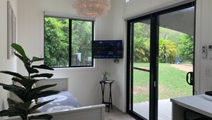 A modern room with a bed, a TV, a small table with a plant, and a sliding glass door to a garden.