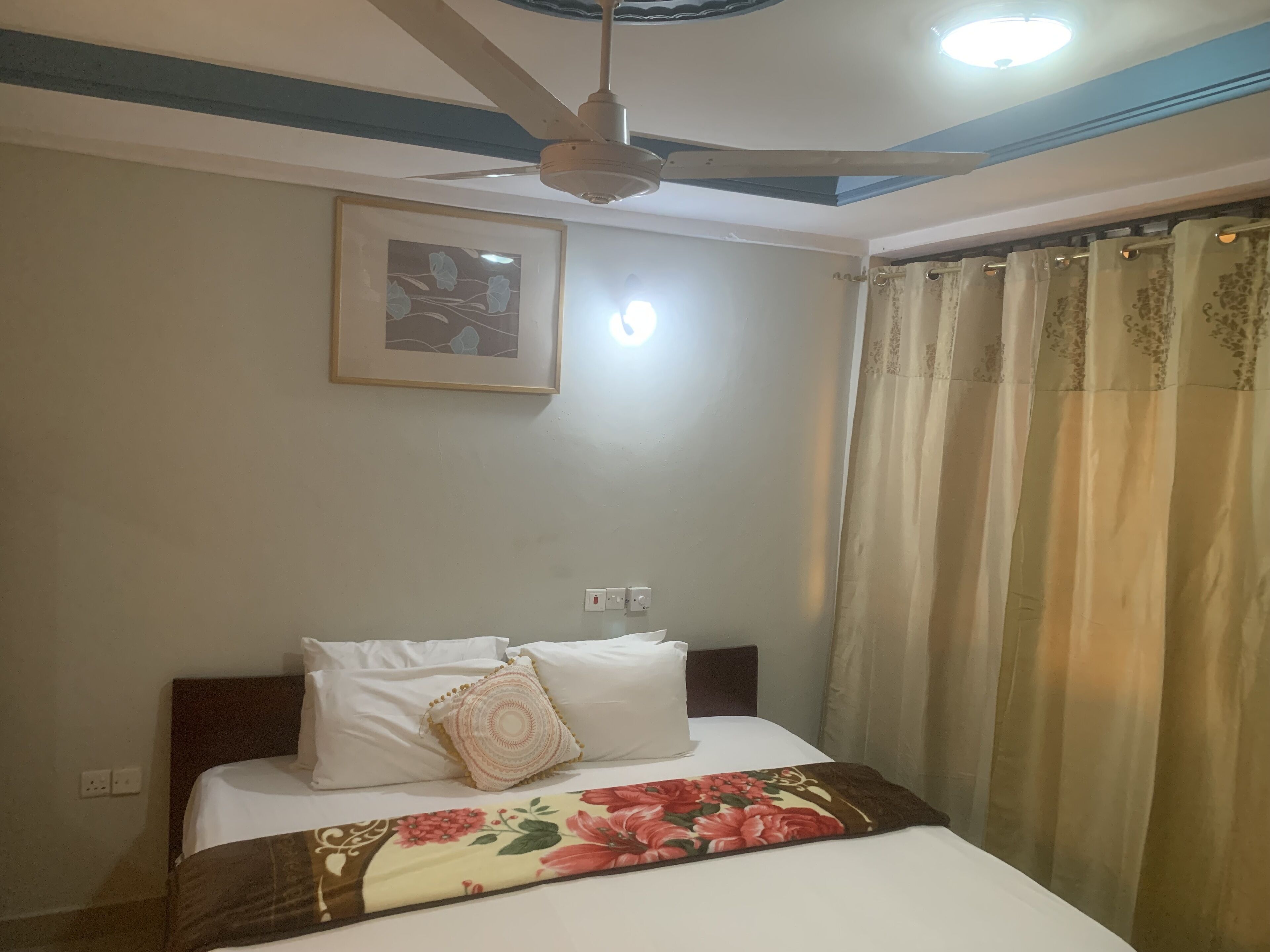 Habitación de hotel con cama, ventilador de techo, un cuadro en la pared y ventana con cortinas.