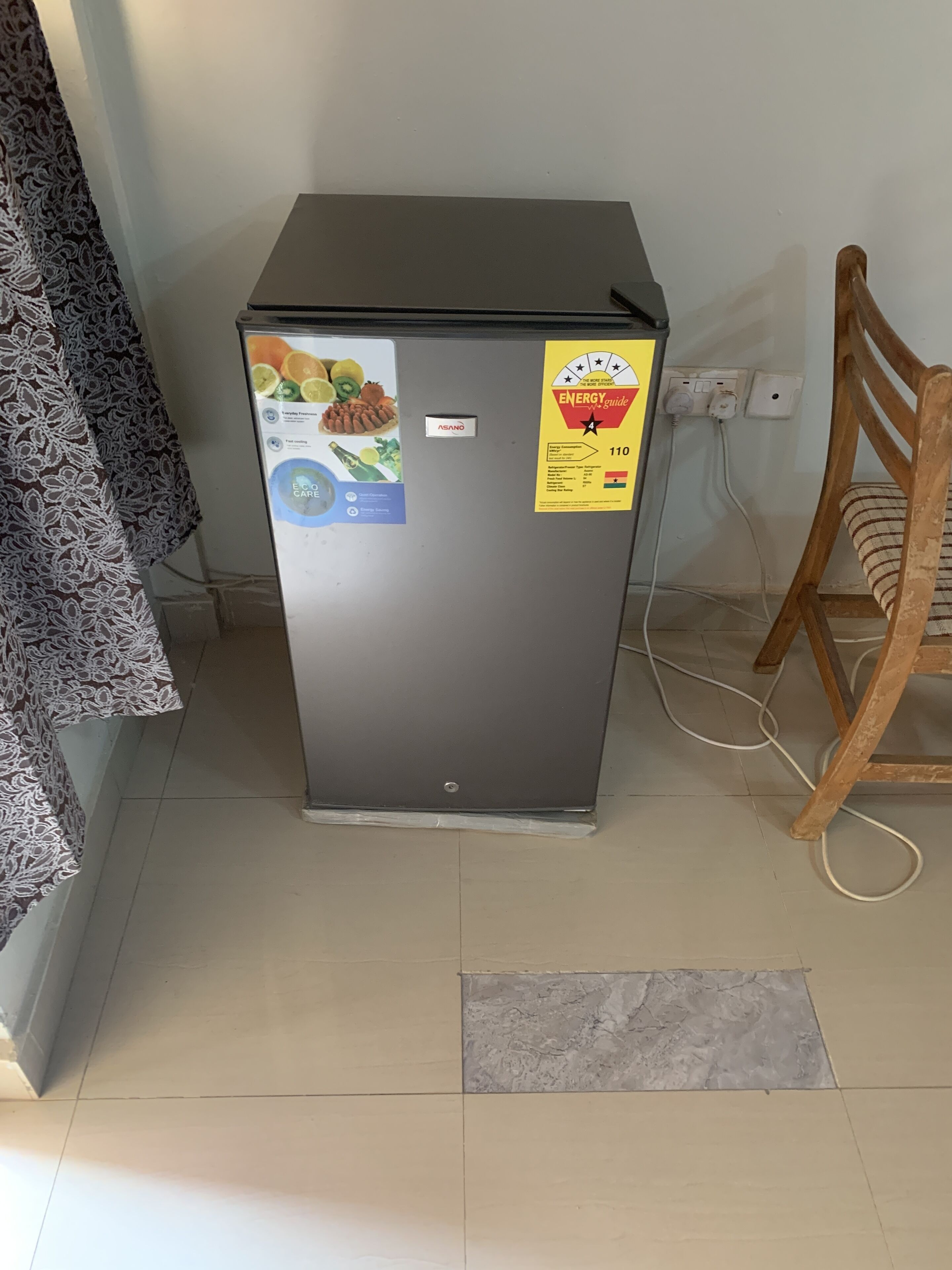 Un petit réfrigérateur doté d’autocollants indiquant son efficacité énergétique, une prise de courant et une chaise en bois à proximité.