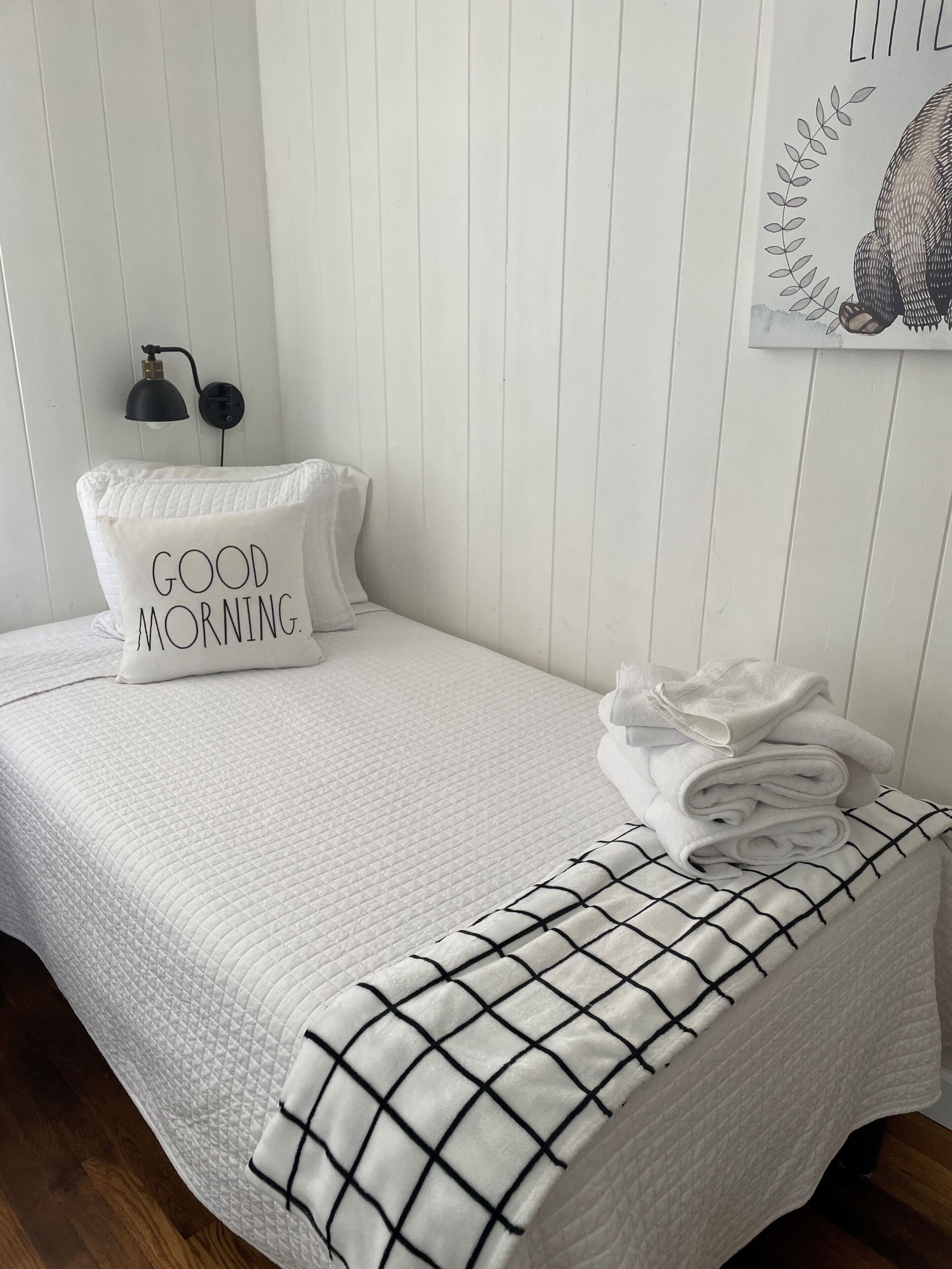 Una cama bien hecha con un edredón blanco acolchado, un almohadón con el texto “BUENOS DÍAS”, una manta blanco y negro a cuadros y una lámpara de pared.