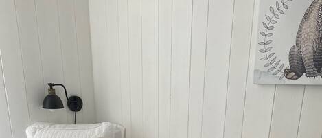 Una cama bien hecha con un edredón blanco acolchado, un almohadón con el texto “BUENOS DÍAS”, una manta blanco y negro a cuadros y una lámpara de pared.