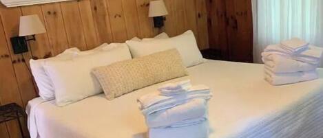 A neatly made bed with white linens and a folded towel on the side, wooden walls, and a small shelf with a plant and picture frame.