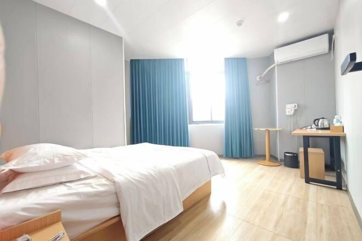 A hotel room with a bed, a desk, a chair, a window with curtains, and an air conditioning unit.