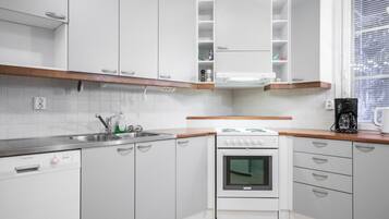 Une cuisine avec des meubles blancs, une cuisinière, une machine à café et une fenêtre avec des stores.
