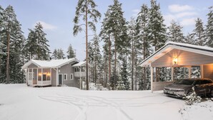 En toetages hytte med en overdækket veranda og en carport, omgivet af sneklædte træer.