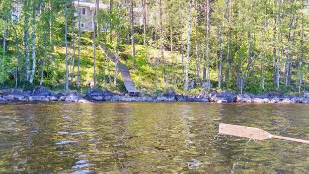 En træpagaj i klart vand med skov og et hus i baggrunden.