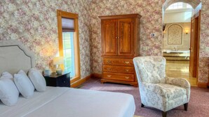 Una camera da letto con un letto, un armadio in legno, una sedia e un bagno visibile attraverso un arco.
