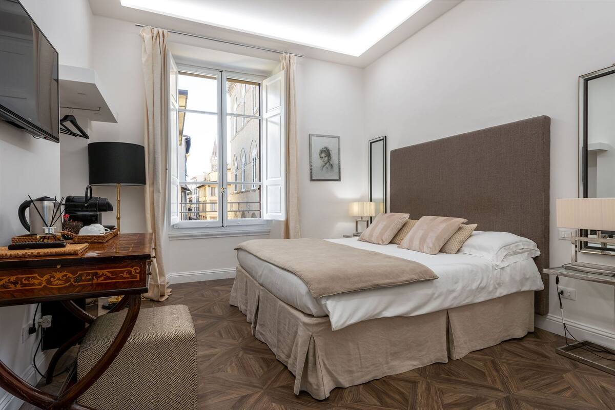 A bedroom with a large bed, a desk with a lamp, a TV, and a window with curtains.