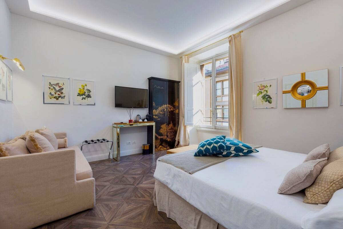 A hotel room with a bed, a sofa, a desk, a TV, and artwork on the walls.