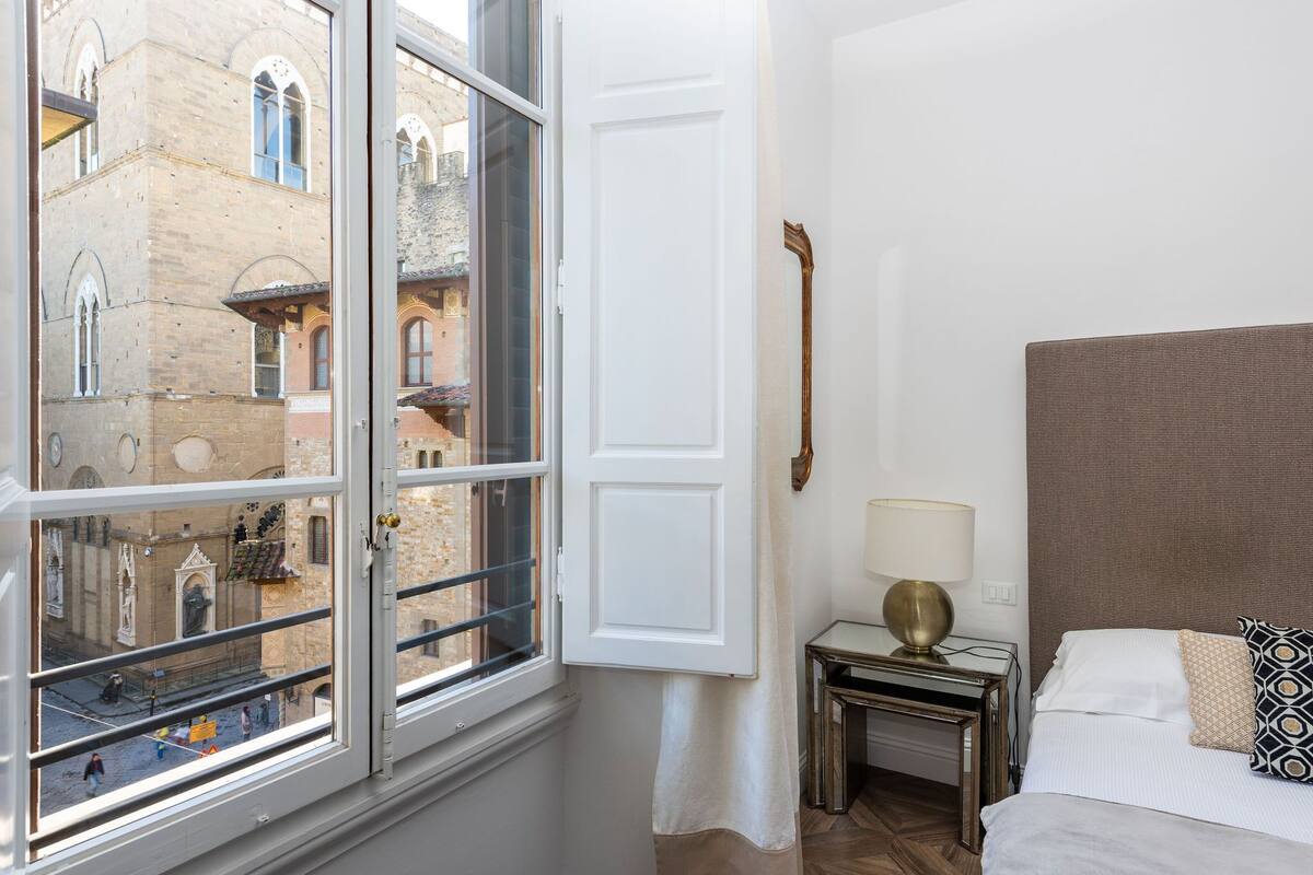 A hotel room with a large window, a bed with a headboard, a bedside table with a lamp, and a view of historic buildings outside.