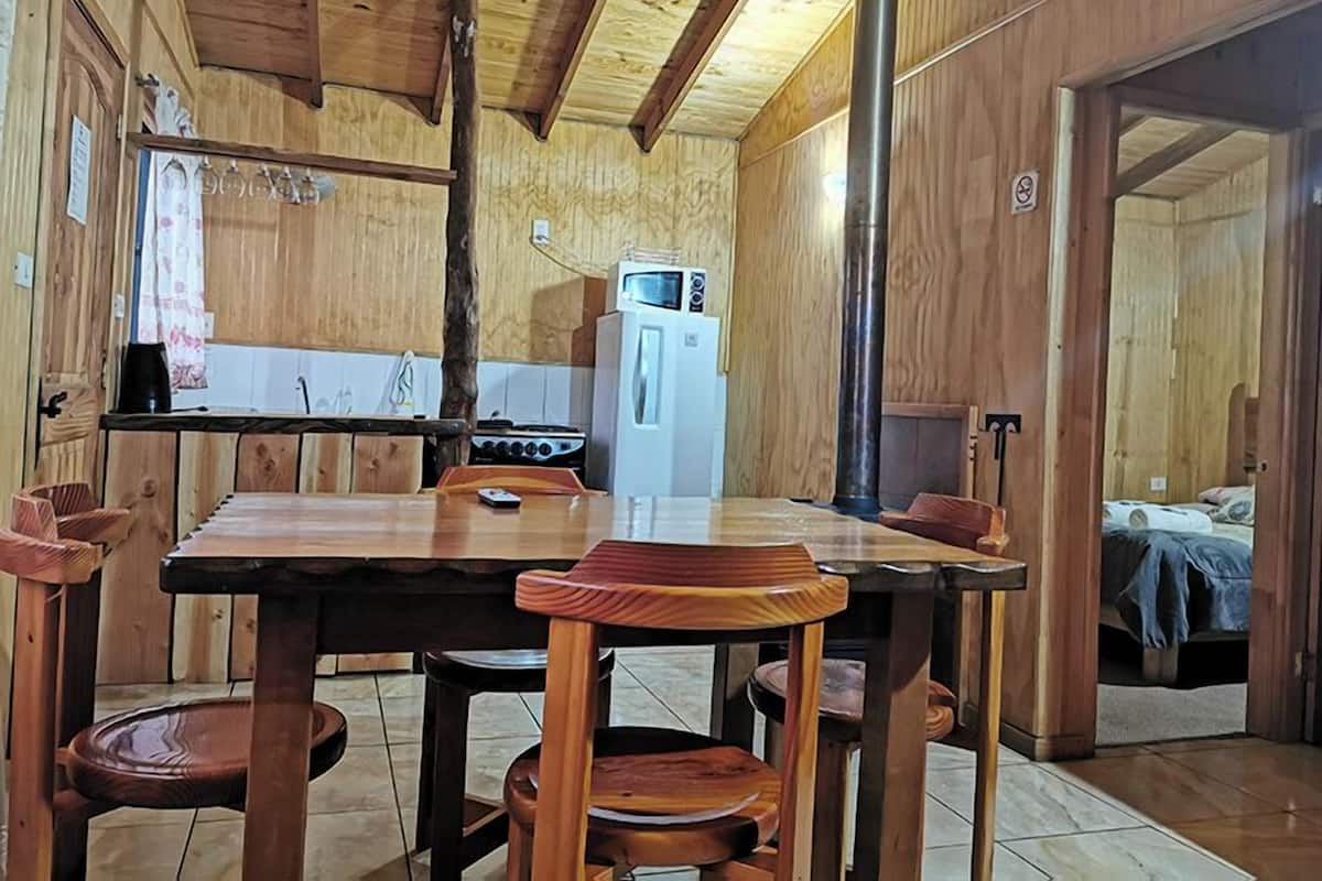 A wooden interior with a dining table, chairs, a kitchen area with a stove and refrigerator, and a bed in the adjacent room.