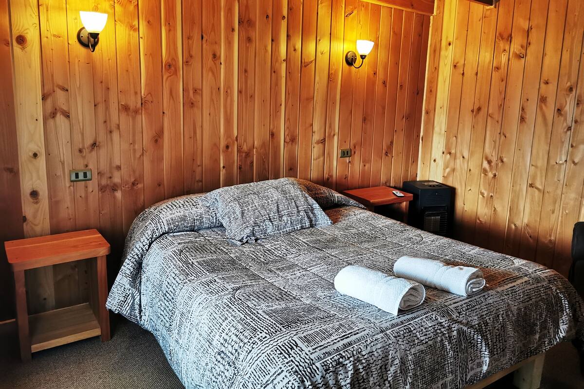 A wooden-paneled room with a bed, two wall-mounted lights, a small table, and a heater.