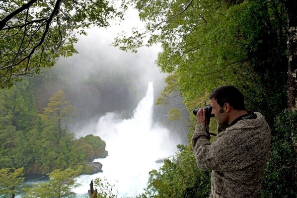 A person taking a photograph of a waterfall surrounded by lush greenery. - Cabañas Choshuenco (Panguipulli)