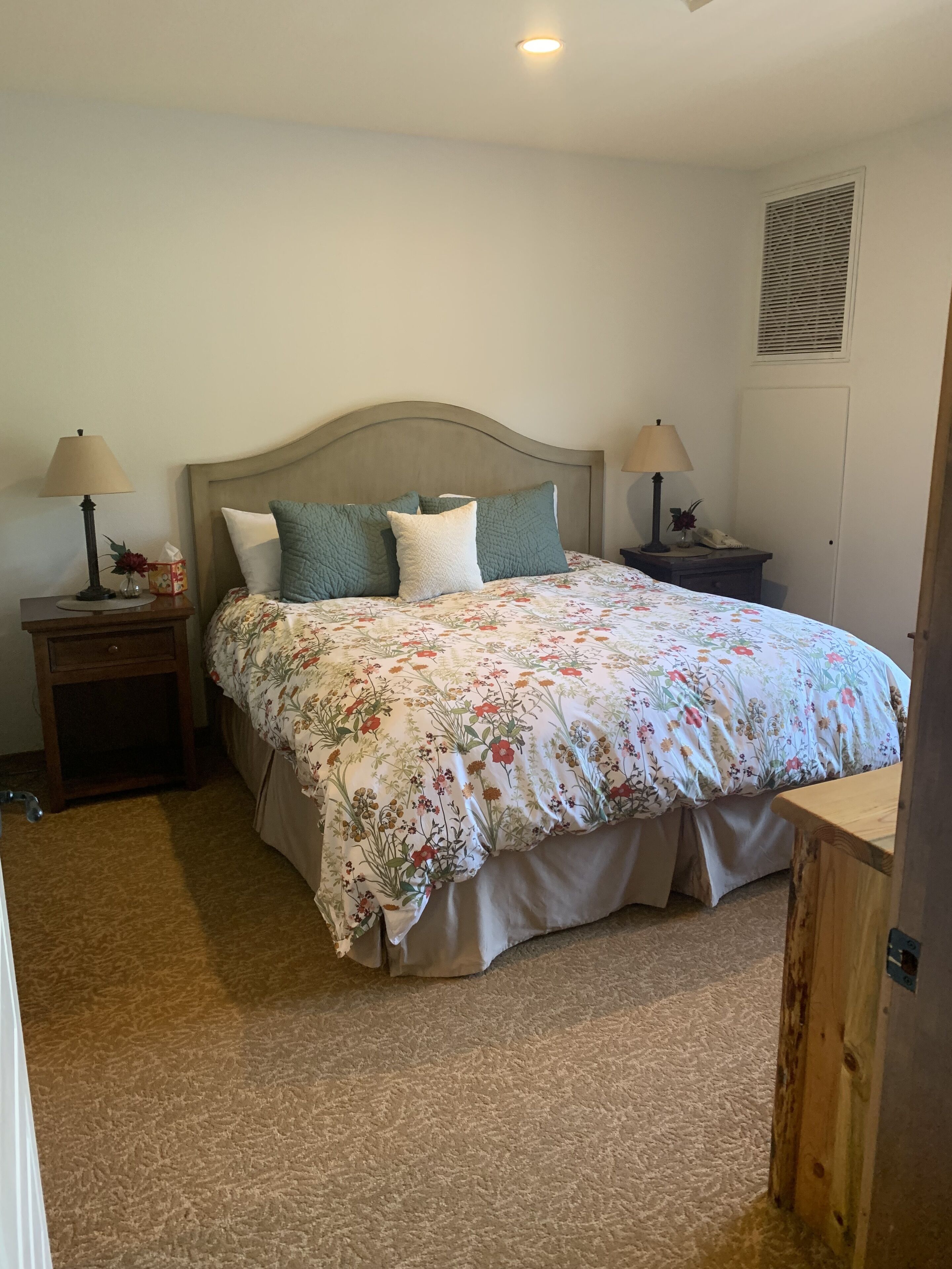 Une chambre à coucher comprenant un lit, deux tables de chevet, une commode, une lampe, une grille d’aération au mur et une porte.