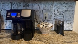 Une machine à café dotée d’un écran numérique, un panier rempli de capsules de café et une cafetière posée sur un plan de travail de cuisine.