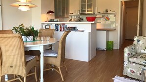A modern kitchen with white cabinetry, a central island with a red bowl, and a dining area with wicker chairs and a glass table. - Cosy, nicely furnished apartment with spacious sunny terrace (Wenningstedt-Braderup (Sylt))