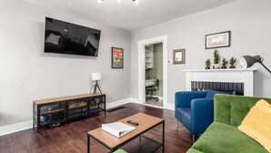 Een moderne woonkamer met een flatscreen televisie, een open haard, groene en blauwe banken, een bijzettafeltje en een boekenkast.