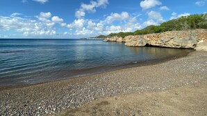 Een kiezelstrand met helderblauw water, rotsachtige kliffen en een verre kustlijn.