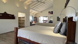 Een slaapkamer met een groot bed, houten hoofdbord en een plafondventilator. In de hoek staat een bureau met stoel, en een raam zorgt voor natuurlijk licht.