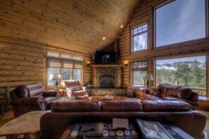 A log cabin interior with a stone fireplace, leather sofas, and large windows offering a view of the outdoors.