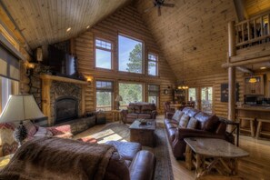 A log cabin interior with a fireplace, leather furniture, large windows, and a stone accent wall.