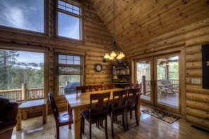 Cozy cabin interior with a dining area, high ceiling, and large window offering a forest view.